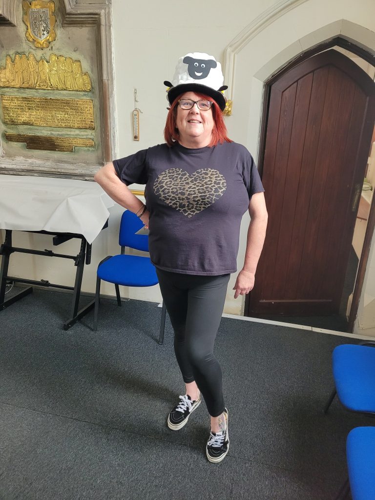 A lady with red hair and glasses wearing a home made sheep hat. She is standing by a door in St Mary's Rawmarsh church.