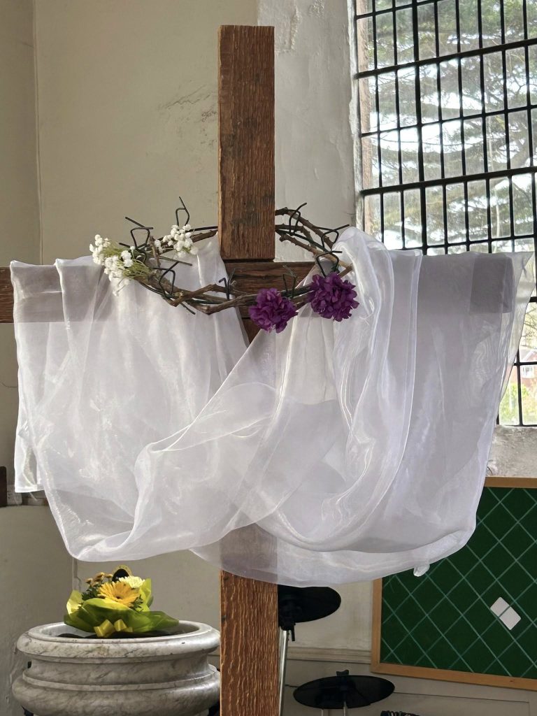 A wooden cross with a cross of thorn and flowers wrapped around it. There is a sheer white clothes over it's arms.