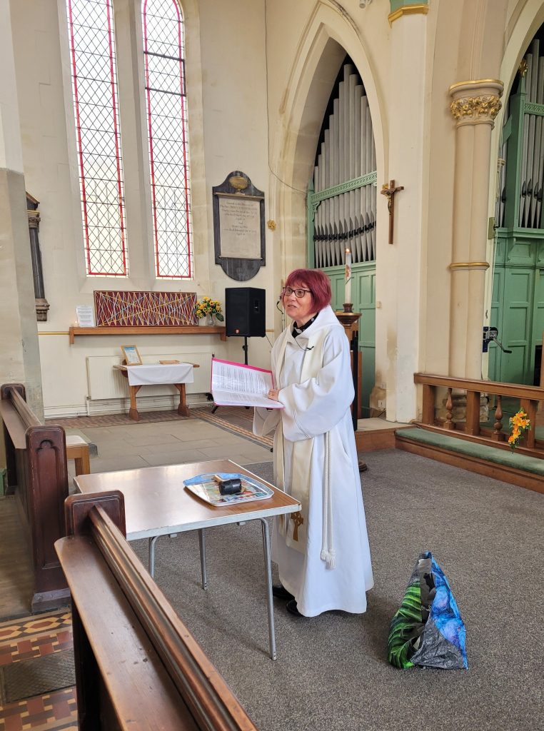 A minister with red hair and glasses wearing ministerial robes stands at the front of St Mary's Rawmarsh church, reading from a red binder