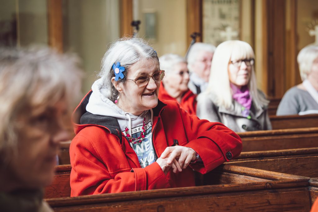 A woman in a red coat with a blue flower in her hair sits in a church pew, laughing