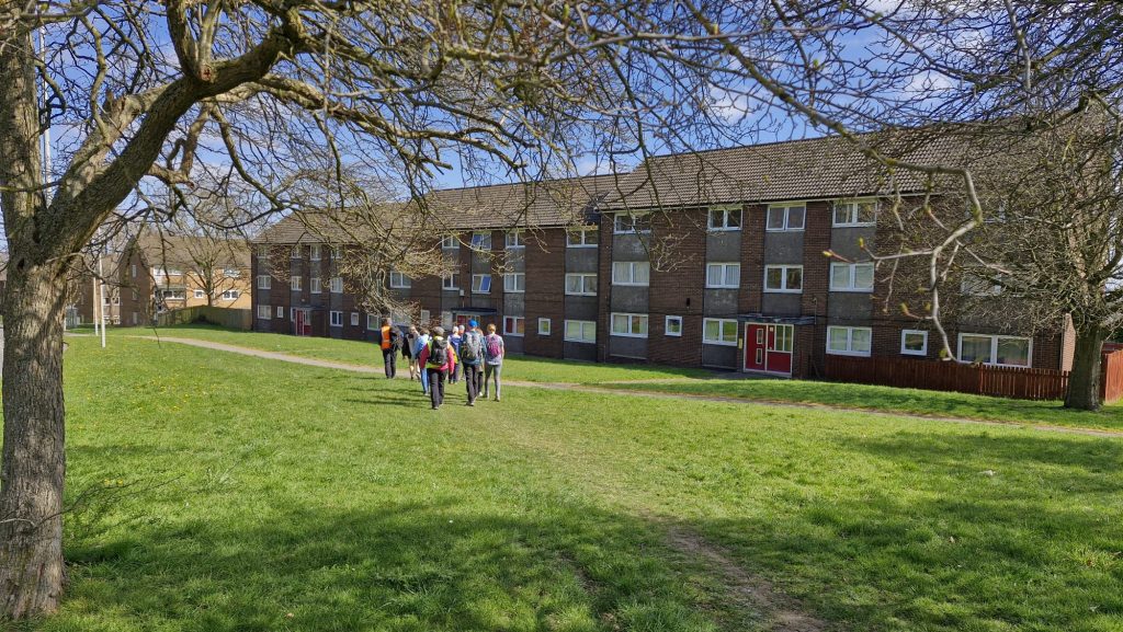A group of walkers walk across a grass field towards a block of 3 story flats