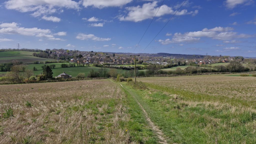 A view of footpath across a field, looking out over a village. There is a pylon in the centre right.