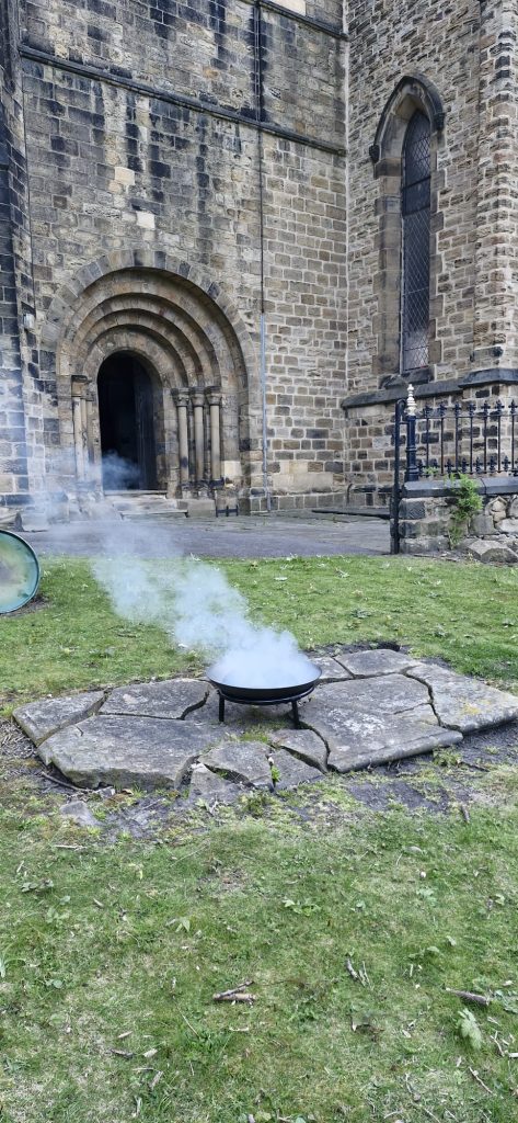 An extinguished fire pit with smoke rising. In the background is the entrance to St Mary's Rawmarsh church.