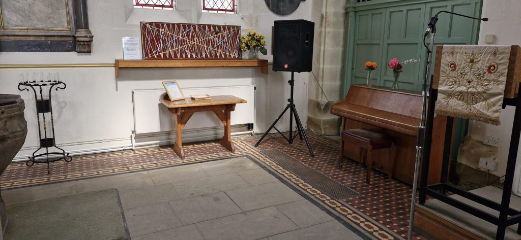 A freshly swept stone floor and tidied corner of St Mary's Rawmarsh church. There is a prayer board with multiple threads of elastic to hold prayers, a small table with candles, a black speaker and a wooden piano in the background.