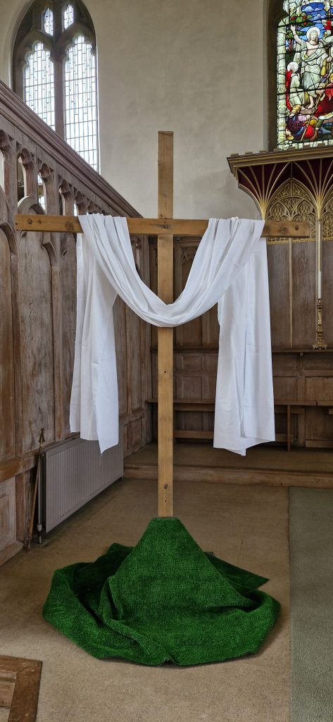 A wooden cross witha white cloth draped over it's arms. At it's base is a green cloth to represent a grass hill. In the background are wooden panels and a stained glass window at St Mary's Greasbrough church.