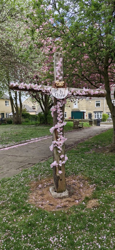 A wooden cross covered with cherry blossoms with the words He is Risen on a board attached to the centre of the cross