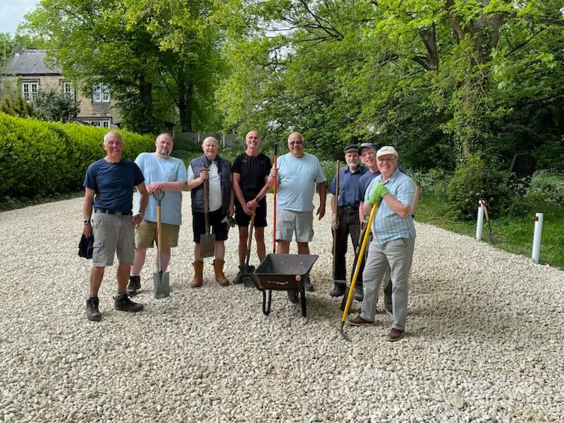 8 men standing on a gravel floor holding spades and a wheelbarrow