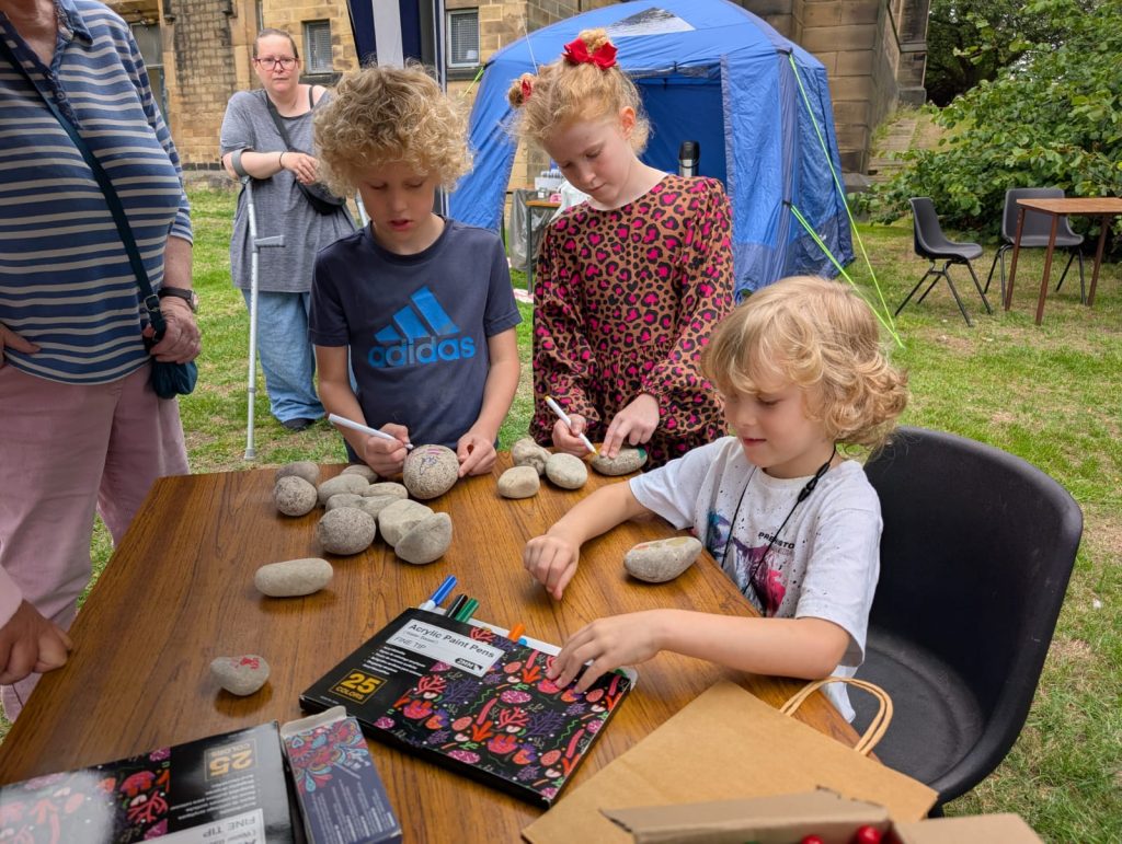 3 children of varying ages stand around a table with rocks on. They are using paint pens to add their own pictures and designs to the rocks.