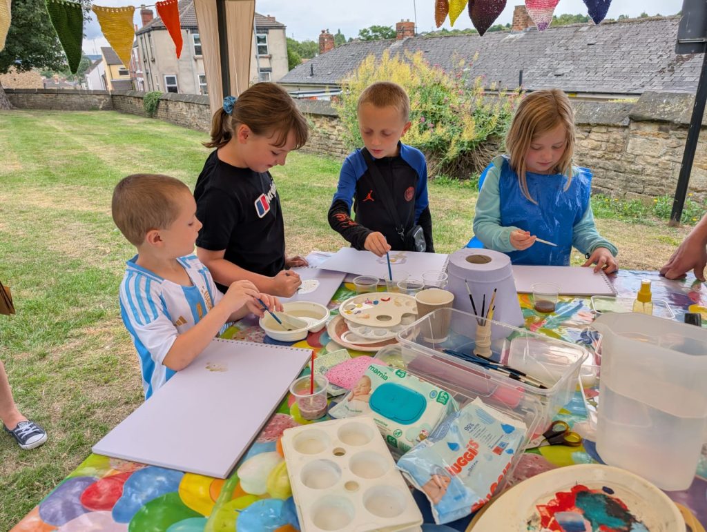 4 children of varying ages stand a round a table full of paper and pain pots. The children are painting pictures of thing in nature using brushes and different coloured paints.