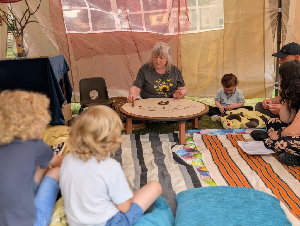 A group of children sit inside a canvas tent, surrounding a table with stones on top that are being used to tell a story by a lady in a grey t-shirt