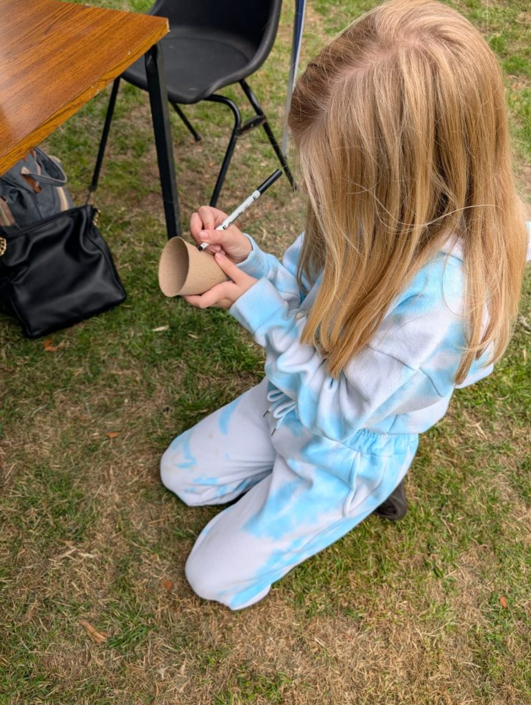 A child decorates a seed box she has planted cress seeds inside. She is sitting on the grass.