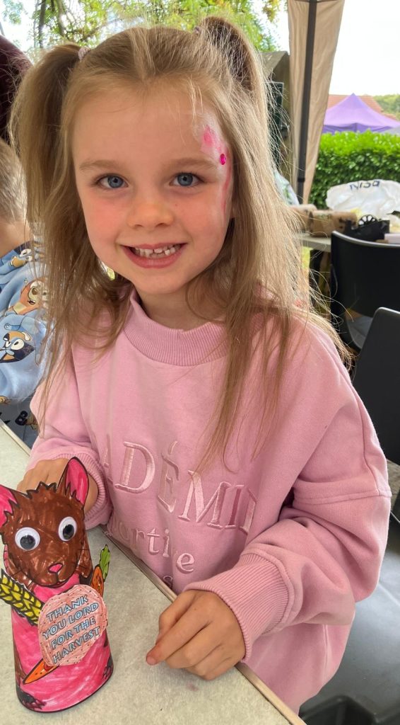Girl child with bunches in her hair holds a paper mouse she has coloured in with felt tips. It is brown and pink.