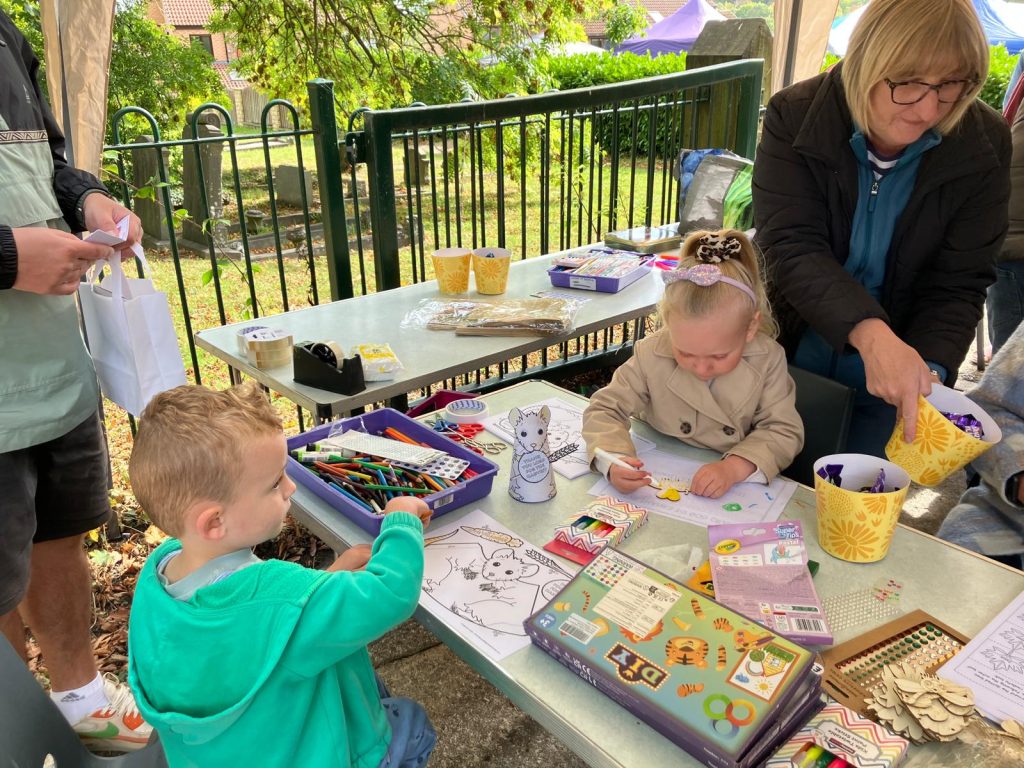 2 Children, a boy and a girl, sit at a table with lots of craft items on it. The girl is colouring her picture of a butterfly whilst the boy looks to his right at a woman giving out chocolates in a yellow bucket with orange flowers.