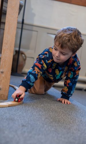 Pre-school boy playing with a wooden train on a wooden track. The train is red and the track is brown. The boy is crawling as he pushes the train along the track. He is wearing a blue jumper with different coloured dinosaurs on. He is crawling on a blue carpet.
